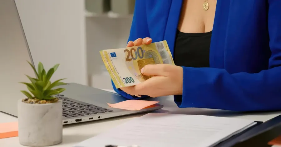 Υπάλληλος με μπλε σακάκι μετράει χαρτονομίσματα ευρώ (200€) δίπλα σε laptop — Κόστος Ασφάλισης Λογιστή