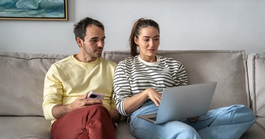 ζευγάρι στον καναπέ συγκρίνει τιμές σε laptop και κινητό — Κόστος ταξιδιωτικής ασφάλισης - Τι καθορίζει την τιμή