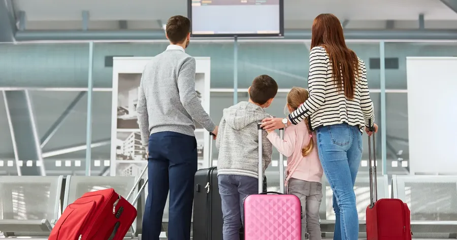 οικογένεια με αποσκευές μπροστά σε οθόνη πληροφοριών αεροδρομίου — Ποιες καλύψεις περιλαμβάνει η ταξιδιωτική ασφάλιση